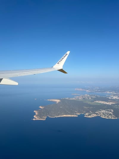 An aerial photograph depicts a view from a Ryanair aircraft, with a portion of its white wing and wingtip, featuring the blue "RYANAIR" logo, visible in the upper left of the frame. The scene below showcases the coastline of Sol de Mallorca, Spain, characterized by a prominent, vegetated peninsula extending into the deep blue Mediterranean Sea. Numerous white and light-colored buildings, consistent with residential or resort developments, are visible scattered along the shorelines and inland areas of the peninsula, particularly clustered in several bays and coves. A smaller island or rock formation is situated off the peninsula's tip, and a tiny white boat is discernible in the water off its western coast. The sky above is clear and bright blue, with a slight atmospheric haze visible on the distant horizon where sea meets sky.