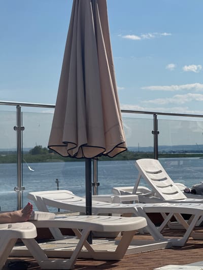 The image shows a waterfront area with a closed, light-colored umbrella dominating the foreground. Several white plastic lounge chairs are arranged on a wooden deck. In the lower left, a person's bare foot with red painted toenails is visible, resting on one of the chairs, with a book placed underneath. Beyond the chairs and umbrella, a body of water is visible, with a distant shoreline featuring greenery and structures. A glass railing with metal supports separates the deck from the water. The sky is blue with scattered white clouds. The location appears to be a resort or public waterfront area.