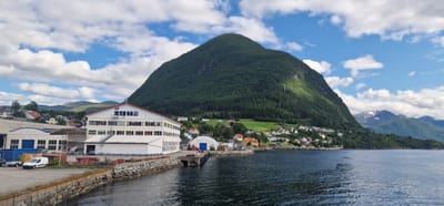 The image captures a waterfront industrial area in Volda, Norway, with a prominent, steep, tree-covered mountain dominating the background. In the foreground, a calm, dark blue body of water laps against a stone embankment and a pier. Several industrial buildings with white siding and red roofs are visible along the waterfront. One large, multi-story building has numerous windows. In the lot next to the buildings, a white van, a trailer, and some large pipes are present. A smaller, rounded blue-roofed building is situated on a pier extending into the water, with a red forklift nearby. Further back, a cluster of residential houses with various colored roofs are scattered on the lower slopes of the mountain. The sky is a bright blue with scattered white clouds. The scene appears to be a working port or industrial zone on the edge of a residential community, nestled against a dramatic natural landscape.