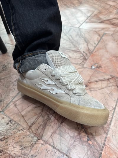 A close-up, low-angle shot shows a person's left foot wearing a light-colored sneaker with white flame accents on the side and a translucent gum sole. The sneaker is laced with thick white laces. The person is wearing dark wash denim jeans that are rolled up at the cuff. The sneaker and jeans are positioned on a floor tiled with marble that has a pinkish-brown and gray veined pattern. The scene is set indoors, likely in a residential or commercial space, given the flooring. There is no specific visual information in the image to pinpoint the location to El Paso, United States of America.