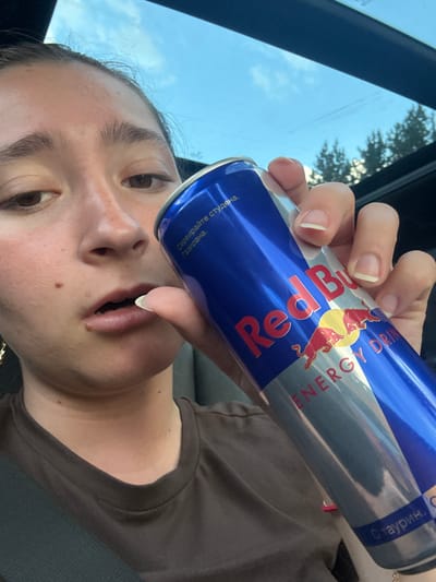 A close-up, selfie-style shot shows a young person's face and a hand holding a Red Bull energy drink can. The person is looking towards the camera with their mouth slightly open. The Red Bull can is predominantly blue and silver, with the brand's logo and text visible. The background suggests an outdoor setting, possibly a car interior with a sunroof showing the sky and trees. The image is not set in Dolno Varshilo, Bulgaria.