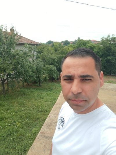 A man takes a selfie outdoors in Bukovlak, Bulgaria. He is wearing a white t-shirt with a black logo on the left chest. The logo appears to be a stylized crest with some text. He has short, dark hair and dark eyebrows. His expression is neutral.
The setting is a rural or semi-rural area with trees and a house visible in the background. The house has a red tiled roof and a brick chimney. There is a grassy area with some small white flowers in front of the trees. A concrete wall runs along the bottom right of the frame. The sky is overcast, suggesting it may be a cloudy day. The time of day is difficult to determine due to the overcast conditions, but it appears to be daytime. There is no specific activity or event taking place; it is a casual portrait.