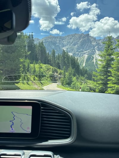 The image is taken from the interior of a car driving on a mountain road. The dashboard is visible in the foreground, with a navigation screen displaying a winding route. The windshield shows a scenic view of a paved road curving through a lush green mountainside. Tall evergreen trees line the road, and in the distance, a majestic mountain range with rocky peaks and patches of snow dominates the horizon under a bright blue sky with scattered white clouds. The location is San Martin de Tor - San Martino in Badia - St. Martin in Thurn, Italy.