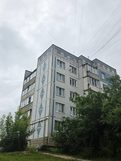 A low-angle, full shot captures a multi-story apartment building on a cloudy day in Mishutino, Russia. The building, with its light-colored tiled facade adorned with a geometric pattern of blue diamonds, dominates the upper portion of the frame. Numerous windows with white frames punctuate the building's exterior. A prominent balcony structure juts out from the middle section. Above and behind the building, a network of thin utility wires stretches across the overcast sky. Lush green trees and bushes partially obscure the lower floors and surround the building's base. In the foreground, a grassy area with a gravel path leads towards the building. No people are visible in the scene.