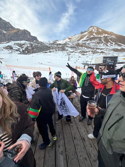 This image captures a lively scene on a wooden deck at a ski resort, likely in Alagna Valsesia, Italy. A group of people, mostly young adults, are gathered, some standing and some seated, enjoying a break from skiing or snowboarding.

In the foreground and midground, several individuals are visible. Many are dressed in winter attire, including ski jackets, hats, and goggles. One person in a dark jacket holds a clear plastic cup with a dark beverage. Another individual wears a black cap with "WEDZE" written on it. Several people are holding or wearing white t-shirts that bear text; a prominent one reads "International Society of the [partially obscured word] University". One person wears a green, yellow, and red striped arm band, possibly representing a flag or country. The overall mood appears relaxed and social.

The background features snow-covered ski slopes and majestic, rocky mountains under a bright sky with some scattered clouds. Ski lift mechanisms and orange safety netting are visible on the slopes. A building, possibly a ski lift station or a mountain restaurant, stands further up the slope, with the word "ALAGNA" and "VALSESIA" visible on its side. The weather appears clear and crisp, typical for a winter day in the mountains. The wooden deck surface is visible in the lower right and mid-right parts of the image.