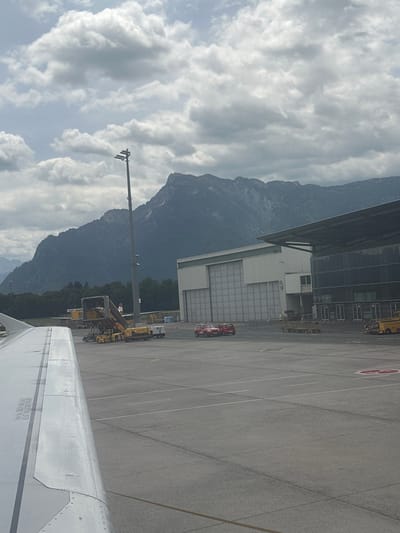 The image depicts an airport tarmac with a mountain range in the background. In the foreground, the wing of an airplane is visible on the left. The tarmac is paved with asphalt and marked with white lines. Several airport vehicles, including baggage tugs and stairs, are present. Two red cars are parked on the tarmac. In the middle ground, a large building, possibly a hangar or terminal, is visible. The sky is cloudy. The location is identified as the airport in Viehhausen, Austria.