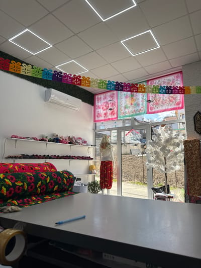 This image presents the interior of a retail shop located in Lazhnitsa, Bulgaria. The foreground features a gray counter with a blue pen positioned on its surface and a roll of brown packing tape partially visible on the left edge. Behind the counter, a white wall on the left displays multiple rows of colorful slippers or small shoes on white shelves. Below these shelves, two large rolls of patterned fabric, one predominantly green with red flowers and another partially visible in pink, are stacked.

Further back, near a glass entrance door and window, a white mannequin stands, attired in red patterned pants and a floral headscarf. To the mannequin's right, a white artificial tree with bare branches is visible. Through the glass, an exterior scene includes unpaved ground, sparse vegetation, and distant structures under natural light. Across the top section of the window and door, several colorful, floral-patterned scarves or textiles are suspended as decoration.

The shop's ceiling consists of a grid of white tiles fitted with multiple square LED light panels. An air conditioning unit is mounted on the upper part of the white wall above the display shelves. A festive garland composed of multicolored gift-box shapes hangs horizontally across the upper portion of the room, situated beneath a strip of green artificial foliage that adorns the wall. No individuals are present in the depicted scene.