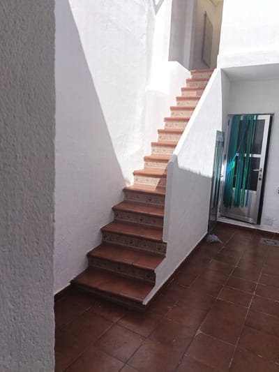 The image depicts an outdoor staircase with terracotta tiled steps in Campillo de Altobuey, Spain. The staircase leads upwards to a doorway with a white frame, adorned with teal and green fabric strips hanging as a curtain. The walls are white and textured, with strong sunlight casting dramatic shadows across them. The ground is also tiled with reddish-brown square tiles. A mop with a blue string head rests on the floor near the doorway. The scene suggests a sunny day, likely midday, given the intensity of the sunlight and the sharp shadows. The setting appears to be a residential courtyard or a patio area. There is no visible text in the image.
