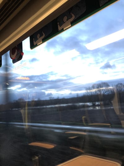 A window seat view from a train, showing blurred scenery in motion. The sky is bright with white clouds. Trees and buildings pass by, reflected in the window. Inside the train, overhead compartments are visible with icons indicating seating. A red emergency handle is also visible, with text in French. The overall impression is one of travel and speed.