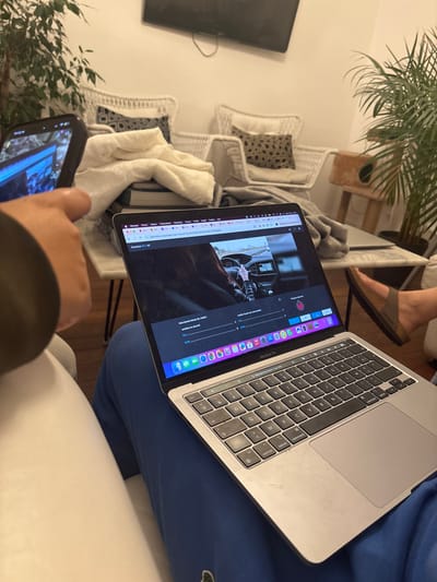The image portrays an indoor living room scene located in Maisons-Alfort, France. In the foreground, a person's right hand holds a black smartphone with a bright screen displaying an image or video. Immediately to the right, a silver MacBook Pro laptop rests on the lap of another individual, who is wearing blue sweatpants featuring a green crocodile logo. The laptop screen displays a web browser with a French driving theory question ("Une bonne tenue du volant") and an image of a person's hands on a car steering wheel, alongside multiple-choice options and the macOS Dock. The right foot of the person operating the laptop, clad in a brown Birkenstock-style sandal, is visible extending from the lower right.
In the midground, a white marble-top coffee table with dark, slender metal legs holds a stack of books partially covered by a cream-colored blanket. The background reveals a white wall and two white wicker armchairs, one with a leopard print pillow, suggesting a seating arrangement. Above the chairs, a dark, wall-mounted flat-screen television is visible. To the far right, a portion of a light-colored wooden cat tree and a large green potted plant are present. Another large green plant frames the left side of the background. The floor is dark, and the overall setting is a furnished residential living space.