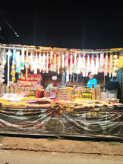 In Bagerhat, Bangladesh, a night market stall displays a variety of goods. The stall is illuminated by overhead lights, casting a warm glow on the merchandise. Hanging from the ceiling are numerous decorative strings of beads and tassels in various colors, including white, blue, green, yellow, and pink.

In the background, two individuals are visible. On the right, a young person wearing a blue t-shirt stands behind the counter. To the left of the young person, an older individual wearing a white cap and a light-colored tunic is seated.

The stall itself is laden with products, primarily packaged goods. There are stacks of small yellow containers, likely containing spices or food items, arranged in neat rows. Larger packages with red and yellow branding are also visible. In the foreground, the stall is covered by a draped fabric with a printed pattern, partially obscuring the lower shelves. The ground in front of the stall appears to be dirt and gravel, with some sparse vegetation.