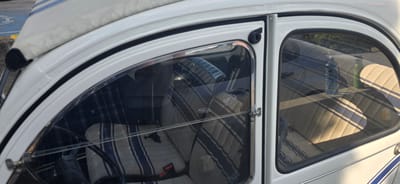 The image shows a close-up of the interior and exterior of a white vintage car, possibly a Citroën 2CV, parked outdoors. The car has a canvas roof with blue stripes. The interior features upholstered seats with a distinct pattern of beige and blue stripes. A black steering wheel is visible. On the passenger side, a blue plastic water bottle and a green water bottle are placed on the seat, visible through the window. The car appears to be parked on asphalt, with a hint of a blue painted area, possibly a parking space, in the background. The lighting suggests it is daytime, with sunlight casting shadows on the car's interior. The image was taken in El Bruc Residencial, Spain.