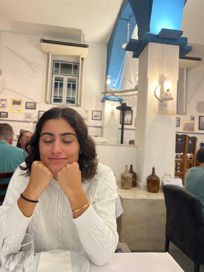 The image shows the interior of a restaurant in Coruche, Portugal. A female individual is positioned centrally in the foreground, seated at a table. She has dark, wavy, shoulder-length hair, her eyes are closed, and a faint smile is present. Her hands are clasped together, resting beneath her chin. She wears a white cable-knit sweater, a black beaded bracelet on her left wrist, and a thin gold-colored bangle on her right wrist. On the table directly in front of her, a white placemat, a folded white napkin, and an empty clear glass are visible.
The background reveals the restaurant's decor, characterized by white walls adorned with various framed pictures and a decorative white relief of a fork and spoon. A blue and white tiled dado rail runs along the lower section of the walls. An air conditioning unit is mounted above a white and blue-trimmed interior window frame. The ceiling features a prominent light blue arched section and structural elements. A large, nautical-themed light fixture, composed of light-colored branches with integrated lantern-style lights, hangs from the ceiling. Additional lantern-style wall lights are affixed to pillars and walls throughout the space. In the mid-ground, a counter displays several large, antique-style glass bottles, some containing preserved food items. Other patrons are partially visible in the background; a man in a teal shirt is to the left, and another person in a similar shirt is to the right, seated at separate tables. The lighting within the restaurant is warm.