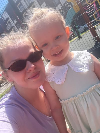 A close-up, selfie-style shot features a woman and a young child outdoors on a sunny day. The woman, in the foreground on the left, wears a purple t-shirt and dark, cat-eye sunglasses. She has her hair pulled back and is wearing a nose ring and small hoop earrings. To her right, the child is looking at the camera with a wide smile, wearing a light green dress with a white lace collar. Behind them, a playground structure with colorful elements is visible, along with a modern building and a chain-link fence. The overall impression is a casual, happy moment captured in an urban outdoor setting. It is not possible to determine the specific location within the city of Vyazniki, Russia, from the visual information provided.