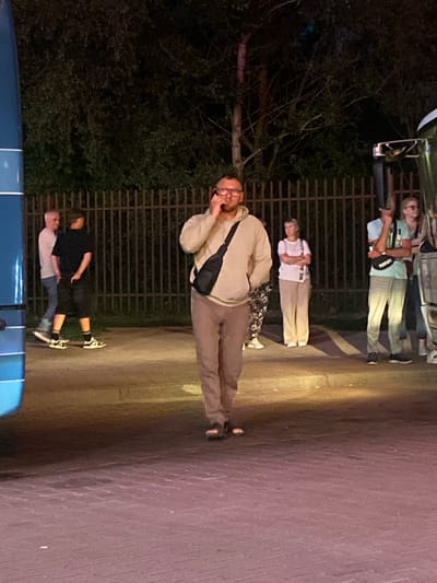 A group of people are standing and walking near a bus at night in Terespol, Poland. The main subject, a man wearing glasses and a light brown hoodie, is walking and talking on his phone. He has a black fanny pack across his chest. Behind him, a metal fence lines the background. Other individuals are visible, including a man in a black t-shirt and shorts, and several people standing near the bus, some of whom appear to be passengers. The ground is paved with reddish bricks. The lighting suggests it is nighttime, with some artificial illumination casting shadows.