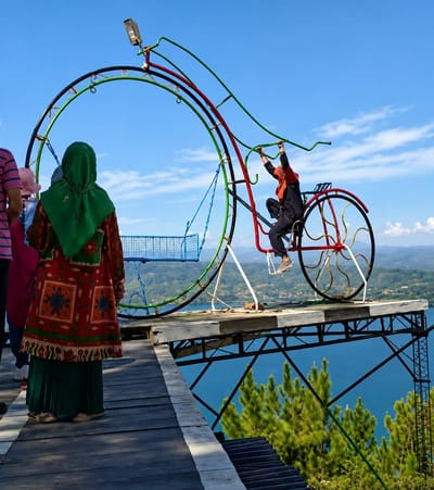The image depicts a scenic overlook in Parik Sabungan, Indonesia, featuring a large, colorful, bicycle-themed amusement ride against a backdrop of lush green hills and a vast blue lake under a clear sky. A person in dark clothing and a headscarf is seated on the bicycle, holding onto its handlebars. In the foreground, three individuals are positioned on a wooden walkway. One person, wearing a green headscarf and an intricately patterned red and green garment, faces away from the camera, looking towards the ride and the expansive landscape. Another person, partially visible on the left, wears a striped shirt. To their right, a third person is wearing a pink hat. The amusement ride is constructed with metal tubing painted in green, red, and black, forming a large circular structure with a bicycle frame integrated into its design. A small blue netted basket is suspended from the circular frame. The wooden walkway extends towards the edge of a cliff, offering a dramatic view of the lake and the surrounding terrain, with a cluster of pine trees in the lower right corner of the frame. The overall scene suggests a tourist attraction or recreational area.