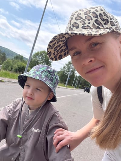 A medium shot captures a woman and a young child in a selfie-style photograph. The woman, positioned on the right, wears a leopard-print baseball cap and a white t-shirt. She is looking at the camera with a slight smile and has her right hand resting on the child's shoulder. The child, on the left, is wearing a dark bucket hat with a green leaf pattern and a brown jacket with the word "Smile" embroidered on it in white. The child is looking to the left with a neutral expression.

The background suggests an outdoor setting, likely on the side of a paved road in the city of Kurdur, Russia. A light pole is visible behind the child, and trees and a gentle hillside are in the distance. The sky is cloudy. A dark car is driving away on the road in the background. The lighting indicates it is daytime, possibly a slightly overcast day.