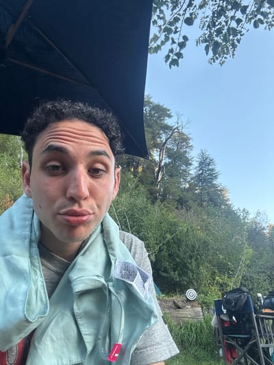A curly-haired person poses for a selfie outdoors, tilted on their side. They wear a light blue garment. Overhead, a partial dark umbrella is visible against a bright sky. Trees and greenery fill the background, and a small target is mounted on a wooden post. A metal-framed chair with bags sits in the top right corner. The selfie focuses on the person's face, capturing a relaxed, playful expression.
