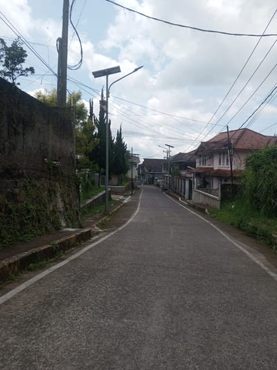 The photograph depicts a paved road winding through a residential area. The road surface is dark grey and textured, with a faint white line marking the edge. On the left side of the road is a high, grey stone wall covered in vegetation. A low curb runs along the base of the wall. On the right side, houses with red tile roofs line the street. One house is painted pink with white window frames. Lush green foliage grows along the roadside. Overhead, a network of electrical wires crisscrosses the sky. A solar-powered street lamp stands on the left side of the road. The sky is overcast with patches of blue visible. The scene is in a quiet, likely residential part of Ban Phouviengnoy, Laos. No people are visible in the immediate foreground.