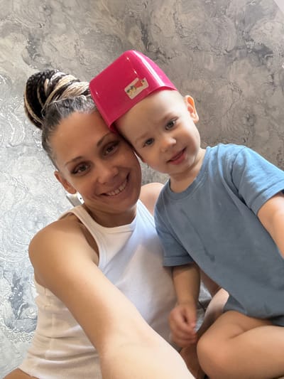 A woman and a child are in a selfie-style photograph. The woman is wearing a white tank top and has her hair up in braids. The child is wearing a blue shirt and has a pink plastic object on his head. They are both smiling at the camera. The background appears to be a textured wall. The location is not discernible from the image alone. I cannot determine the city of Yaroslavl', Russia as a location.