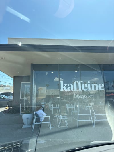 Here's a forensic description of the image:

**Location:** Redondo Beach, California, United States of America.

**Subject:** A storefront for "Kaffeine Espresso Bar".

**Exterior:**
*   The building has a modern architectural style with a flat roof, grey wall finish.
*   A glass storefront is prominently displayed with the business name "Kaffeine" vertically oriented in white letters, along with "Espresso Bar" printed beside the business name.
*   Through the glass, white outdoor seating can be seen, which includes tables and chairs.
*   Reflections on the glass indicate the presence of traffic, other businesses, and some greenery across the street.

**Interior:**
*   The interior is visible through the glass, suggesting a cafe-style setup with chairs and tables.
*   Patrons are seated and appear to be engaged in conversation.

**People:**
*   Individuals can be seen seated at tables through the glass window, inside the café.

**Objects:**
*   Cars are reflected in the glass.
*   The business name "Kaffeine" in large, white letters.
*   White cafe-style chairs and tables.
*   Exterior plants.

**Environment:**
*   The environment is urban, suggesting a business district or street with pedestrian activity.
*   The daylight indicates a sunny day.

**Inferences:**
*   The storefront belongs to a coffee shop named "Kaffeine Espresso Bar".
*   It is an active business with patrons inside.
*   The business appears to have outdoor seating.