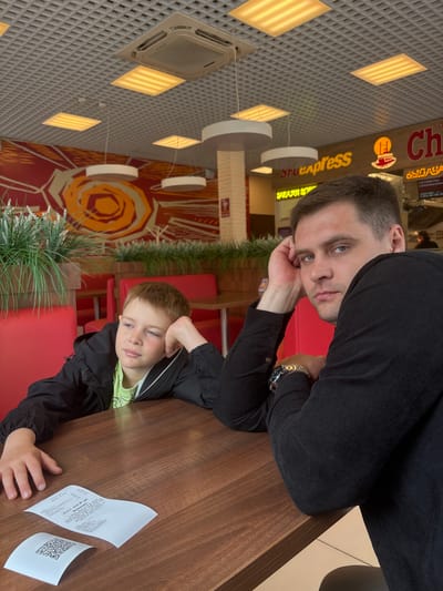 Inside a food court or casual dining establishment in Krilovo, Russia, an adult male and a boy are seated at a dark brown wooden table. The adult male, approximately 20-30 years old with short brown hair and blue eyes, wears a black long-sleeved shirt and a metallic watch on his left wrist. He leans his right elbow on the table, supporting his head with his hand, and looks directly at the camera with a neutral expression. To his left, a boy, approximately 8-10 years old with reddish-brown hair and light eyes, wears a black jacket over a light green t-shirt. He leans his left elbow on the table, supporting his head with his hand, and gazes to the left of the frame with a pensive or bored expression.

On the table in front of the boy are two white paper receipts; one is folded to reveal a QR code, and the other is unfolded, displaying Cyrillic text that includes "Итого: 1519,00 руб." (Total: 1519.00 rubles) and "Внимание!" (Attention!). The background features red upholstered booths and tables, along with decorative planters containing artificial greenery separating seating areas. A large abstract mural in red and orange tones adorns a back wall. Commercial signs in Cyrillic script are visible, including "Snack Express" in yellow lettering and another sign with text partially visible as "Булача" (likely "Bakery"). The ceiling is a gray grid-patterned false ceiling with recessed square lights emitting warm yellow light, an air conditioning unit, and several modern white circular pendant lamps hanging down.