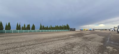 A wide, cracked asphalt lot is the foreground of this outdoor scene, with a row of tall, dark green cypress trees lining a green metal fence in the middle ground. Behind the trees, a line of wind turbines is visible in the distance against a stormy, bruised purple sky.

Several semi-trucks are parked in the lot, some with trailers. On the left, a black truck with "Martín Cargo" printed on its side is parked. Further to the right, a line of trucks are visible: a red and blue truck, followed by two yellow trucks, one with "DISCORDIA" written on its trailer, and then a white truck. On the far right, a white truck is parked, and another truck is partially visible.

Streetlights are scattered throughout the lot, some of which are illuminated. The time of day appears to be late afternoon or early evening, given the dramatic cloud formations and lighting. The weather is overcast and possibly rainy, adding a somewhat somber mood to the scene. The location is implied to be a truck stop or a similar service area, likely in Épila, Spain, given the presence of the wind turbines and the type of landscape. There is no visible activity of people in the scene.