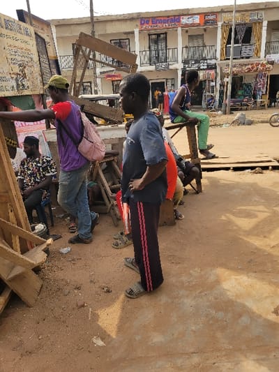 The image depicts an unpaved outdoor urban scene in Puerto López, Colombia, under bright daylight. In the foreground, a dark-skinned male, approximately young adult, stands in profile facing left. He wears a dark blue t-shirt, black track pants with two pink vertical stripes, and grey slides, with his left hand resting on his hip. To his left, another dark-skinned male wears a yellow baseball cap, a purple t-shirt over a red long-sleeved shirt, blue jeans, and carries a light pink backpack. He leans towards a weathered wooden structure, possibly a display stand or signboard. A third dark-skinned male is seated on a dark chair to the left of the purple-shirted individual, wearing a multi-patterned shirt. To the right of the central standing figure, a dark-skinned youth sits on a tall, slender wooden stool, wearing a blue sleeveless top and bright green pants, facing right. The ground in the foreground and mid-ground is reddish-brown dirt, with scattered debris and tire marks. In the background, a multi-story white building with commercial storefronts is visible. The uppermost floor displays a sign reading "De Emerald". Other signs, including "DESTINY", are visible on lower levels. Utility poles and additional individuals are present along the unpaved street extending into the background. Shadows indicate direct sunlight from the upper right.