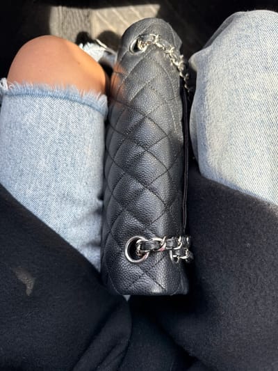 A close-up, high-angle image captures a black quilted handbag resting on a person's lap. The bag features a classic diamond-quilted pattern on its textured black caviar leather surface. A prominent silver-toned chain strap, interwoven with black leather, is visible along the top edges of the bag. The bag is placed on the lap of an individual wearing light blue denim jeans, characterized by their visible fabric weave and casually frayed, distressed hems around the knee. A section of the person's bare skin, likely the knee or thigh, is visible beneath the denim. To the right of the bag and jeans, a dark, soft-textured fabric, possibly a coat or blanket, is present, partially obscured by shadow. The background is dark and out of focus, suggesting an interior environment such as a car or a seated indoor setting. Bright natural light illuminates the scene, creating defined textures and shadows. The image depicts a casual moment in Strömstad, Sweden.