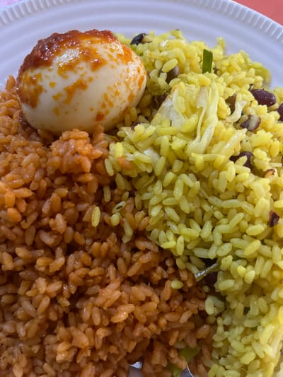 A plate holds two types of rice and a hard-boiled egg. Yellow rice mixed with beans and cabbage sits beside orange rice. The egg, which has a pale yellow shell, is coated in reddish-brown sauce on one side. The plate is white with a ridged edge. The dish appears to be a homemade meal.