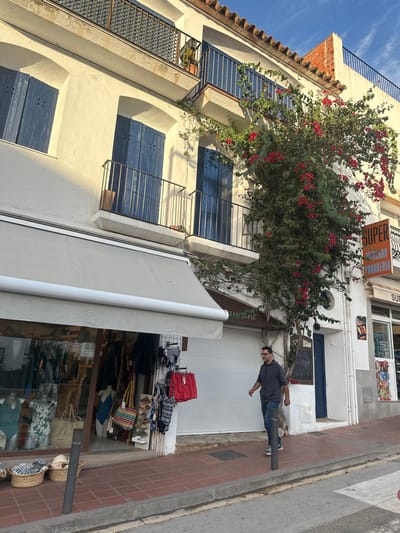 This image depicts a daytime street scene in Calella De Palafrugell, Spain. The foreground features a brick-colored paver sidewalk and a gray asphalt street.
On the left, a two-story building with white stucco walls is visible. The ground floor houses a retail establishment with a large glass display window, showcasing mannequins dressed in swimwear and clothing. Baskets containing merchandise are placed outside, and racks display red shorts and various swimwear. A light-colored retractable awning extends over the storefront. Above, two windows with closed dark blue wooden shutters and dark metal railings on small balconies are visible.
To the right of this, a large, vibrant bougainvillea plant with numerous red-pink flowers and green foliage climbs the white wall of the building, partially obscuring a third window with dark blue shutters and a balcony. Beneath the bougainvillea, a white roller shutter indicates a closed establishment, with the word "restaurant" faintly visible above.
Further right, another storefront is partially visible, featuring a sign with orange text that reads "SUPER MERCADO PANADERIA" (Supermarket Bakery). The upper levels of the buildings feature additional balconies with dark railings and visible terracotta roof tiles. The sky is clear blue with sparse white clouds.
A single male subject is present, walking on the sidewalk from right to left in the mid-ground. He is a middle-aged man with dark hair and glasses, wearing a dark gray long-sleeved shirt or hoodie and blue jeans. He is looking forward and slightly to his left. A dark gray bollard is positioned on the sidewalk to his left. Bright sunlight illuminates the scene, casting shadows.