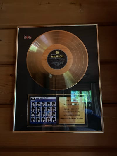 This is a framed gold record by The Beatles for their album "A Hard Day's Night." The record is a light golden color and is mounted on a black matting. Below the record is a smaller framed picture of the album cover and a plaque. The plaque has text that reads: "THE BEATLES," "A HARD DAY'S NIGHT," "SERIAL # RGG-017-17," "IN RECOGNITION OF ITS WORLDWIDE SUCCESS," and "LIMITED EDITION COLLECTORS SERIES." There is also a Union Jack flag in the top left corner of the matting. The frame is silver and hangs on a wooden wall. The background shows a window with greenery outside and some curtains. The image does not show the city of Ленинское, Russia.