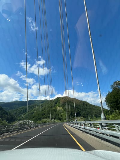 The image captures a view from a vehicle driving across a modern suspension bridge on a clear, sunny day. The bridge has a paved roadway with white and yellow lane markings. Along the sides of the road are metallic guardrails. Numerous vertical cables, characteristic of a suspension bridge, extend upwards into the bright blue sky, which is dotted with fluffy white clouds.
In the background, lush green, tree-covered mountains rise steeply. Some of these mountains show signs of human activity, such as what appear to be power lines or ski lifts traversing their slopes. The bridge itself is set within a mountainous valley.
The location is identified as the bridge over the Psezuapse River in Kepsha, Russia, which is part of the Sochi area. The environment appears to be a natural landscape with significant infrastructure. There are no visible people or animals in the frame, only the man-made structures of the bridge and the natural beauty of the surrounding mountains and sky. The reflections on the car's windshield are also a prominent feature of the image.