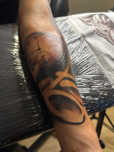 The image shows a close-up view of a person's forearm with a tattoo. The tattoo is a black and orange design that incorporates a clock face with Roman numerals and a smaller dial. The rest of the tattoo consists of abstract, curved shapes in black and orange shading. The forearm is resting on a black surface covered in plastic wrap, which reflects light. In the background, a white sheet with an image of wings is visible. The scene appears to be in a tattoo parlor. The image does not contain any information about the city of Deventer, Netherlands.