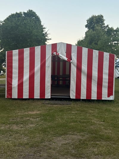 A red and white striped tent stands in a grassy area, possibly a park or fairground, in Завалочыцкі сельскі Савет, Belarus. The tent has an open entrance revealing tables and chairs inside. A white and red striped object hangs from the entrance. Two large trees are visible in the background. The sky is clear and pale.
