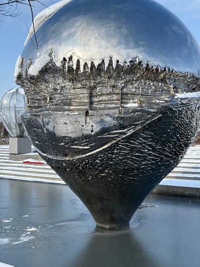 The image depicts a large, metallic, spherical sculpture situated outdoors in what appears to be a public space or park in the city of Plodorodny, Russia, under clear, cold weather conditions. The primary subject is a multi-textured, highly reflective sculpture, predominantly silver in hue. Its upper section is a smooth, polished sphere, partially covered with accumulated snow and ice, reflecting a bright blue sky with diffuse sunlight. Below this, the sculpture transitions into a horizontally segmented, irregular, and roughly textured band, appearing almost molten or organically deformed, also with some snow and ice deposits. Further down, the surface becomes dark and heavily textured with a rippled or hammered pattern before tapering into a smooth, conical base that submerges into a frozen body of water. The ice surrounding the sculpture's base is largely smooth but shows circular patterns and scattered patches of snow. In the background to the left, a second, smaller, spherical sculpture is visible, appearing to be a transparent or translucent globe with a metallic lattice structure, resting on a gray, cubic stone pedestal. Behind these structures, a set of snow-dusted steps or tiered platforms extends horizontally. Bare tree branches are visible against the blue sky in the upper left corner. No individuals or active subjects are present.