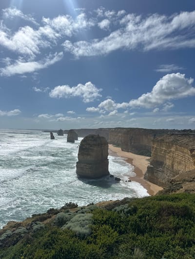 Jagged limestone stacks rise from the ocean along a rugged coastline. Waves crash around the base of the formations. A sandy beach nestles between cliffs. Lush green vegetation covers the clifftop. Above, a blue sky is adorned with wispy white clouds, the sun creating a faint glare. The scene evokes a sense of natural grandeur.