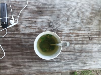 An overhead, eye-level shot captures a white ceramic mug of green tea with leaves and a metal spoon, placed on a weathered wooden table. To the left of the mug, a black portable digital voice recorder and white earbuds with their cord are also on the table. The scene is set outdoors in Шарпілаўскі сельскі Савет, Belarus.