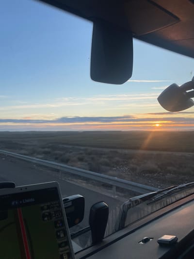A sunrise is captured from inside a vehicle. The sun is visible on the horizon, with bright light radiating from it. The sky is a mix of blue and orange hues. The vehicle's interior, including mirrors and a navigation system, are visible. The landscape outside appears to be a field next to a road. The image is rotated to the side, making the horizon appear vertical.