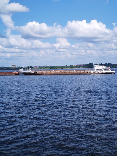 The image captures a wide view of a large, rusty barge being pushed by a white tugboat on a deep blue body of water. The sky above is a bright blue, dotted with fluffy white clouds. In the background, a distant shoreline is visible, featuring a line of trees and several buildings, including what appears to be industrial structures and a church steeple. The water is slightly choppy, indicating a gentle breeze. The scene is devoid of any clear indication of the city of Vasil'ki, Russia, as the background scenery is generic and not specifically identifiable as belonging to that location.