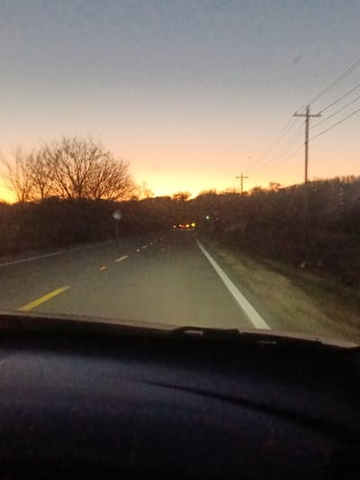 The image depicts a straight asphalt road at either dawn or dusk, viewed from the interior of a vehicle looking forward through the windshield. The sky exhibits a gradient from light blue overhead to prominent orange and yellow hues at the horizon. The two-lane road is marked with a solid white line on the right shoulder and a broken yellow center line, illuminated by the foreground vehicle's headlights. Multiple red taillights from distant vehicles are visible ahead on the road. Dense, dark vegetation, including a prominent bare deciduous tree on the left, borders both sides of the road. Two utility poles with overhead power lines are discernible on the right side of the road. The dark dashboard and the lower frame of the windshield occupy the bottom portion of the image. No individuals are visible. While the requested location is Ozark, United States, specific identifying landmarks within the city are not present in the scene to confirm its precise location.