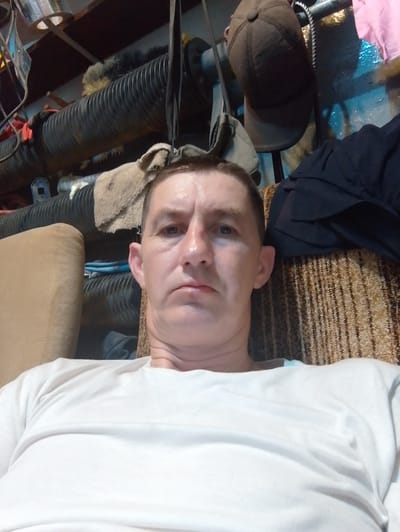 A slightly blurry, medium close-up, point-of-view photograph shows a fair-skinned adult male with short, dark hair and light eyes, looking directly at the camera. He has a neutral expression. He is wearing a plain white t-shirt and is seated in what appears to be a room with various objects in the background. The background includes what looks like industrial piping, a brown baseball cap hanging, and a patterned upholstery. The lighting is somewhat dim and appears to be artificial. The location within the city of Bytom, Poland is not discernible from the visual information provided in the image.
