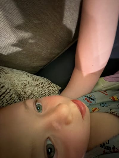 The image is an extreme close-up shot of a young child's face, looking upwards. The child has light skin, green eyes, and reddish-pink lips. A portion of an adult's arm, also with light skin, is visible in the upper right portion of the frame. The child appears to be lying down, possibly on a couch or bed, as indicated by the textures of fabric in the background. One piece of fabric, a patterned throw pillow, is in the lower left. Another piece of fabric, a light blue textile with cartoonish imagery and the words "island trip" printed on it, is visible behind the child's head and arm. The lighting suggests an indoor setting, possibly with natural light coming from the side. The image is very tightly cropped, focusing heavily on the child's features. There is no information within the image to indicate a location in the city of Zmeitsa, Bulgaria.