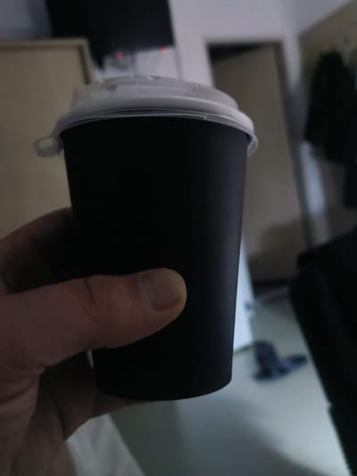 A close-up, blurry, indoor shot shows a hand holding a black disposable coffee cup with a clear plastic lid. The background is out of focus and appears to be a room with a door and a dark object on the floor. The setting is indoors, likely a residential or commercial space, and the image does not provide enough detail to confirm its location in Қосшағыл ауылдық округі, Kazakhstan.