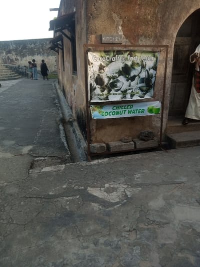 The image depicts an outdoor scene within Fort Jesus, located in the Makadara/Mji Wa Kale ward of Mombasa, Kenya.
In the foreground, a cracked and uneven concrete ground surface occupies the lower half of the frame. To the right, an aged, textured stone wall runs vertically, featuring a narrow, concrete drainage channel at its base. A small, rectangular wooden sign reading "KITCHEN" is mounted on this wall. Below it, a metal stand holds two banners: the upper banner displays "CocoCraft @ Fort Jesus" with images of coconut shell crafts and text reading "Traditional artwork items made from coconut shell"; the lower banner advertises "CHILLED COCONUT WATER" with illustrations of green coconuts. Below these banners, the metal stand forms a shelf holding several rough-hewn concrete blocks and a dark, amorphous object. To the far right, partially visible at an entrance, a person stands wearing a long, light-colored garment and sandals, with a red and black plaid bag hanging over their shoulder.
To the left of the wall, a concrete walkway extends into the midground. Three individuals are visible on this walkway further down: one in light-colored clothing and shorts, another in a striped garment, and a third in dark attire. Their specific actions are indeterminate. Further left, a series of stone steps ascend along another stone wall that forms the outer perimeter of the structure, visible in the background against a light sky. The overall lighting suggests ambient daylight, casting some areas into shadow.