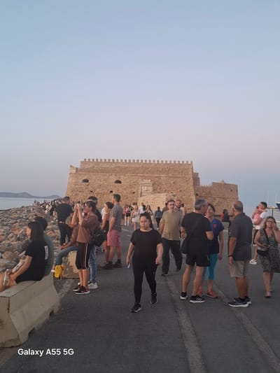 A landscape view depicts a crowd of people on a paved surface near a stone structure with crenellations, possibly part of a fortification or castle, against a backdrop of a light sky.
People are of varying ages and attire, with some appearing to be tourists observing or taking pictures of the scenery. The stone structure is light brown, with distinct architectural details like crenellations and openings that may be windows.
In the background, there appears to be a body of water, potentially the sea, with a hint of land visible in the distance. The paved surface suggests an urban setting and is part of the city of Heraklion Municipal Unit, Greece.