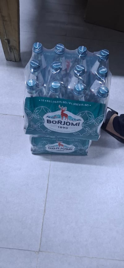 The image shows two stacked packages of Borjomi Georgian natural mineral water bottles. Each package appears to contain 12 bottles, each being 0.5 L (16.9 FL OZ) or 6 L (202.8 FL OZ) in total volume per package. The bottles are clear with blue caps. The packaging is teal and features a deer logo and the brand name "BORJOMI". The packages are wrapped in plastic. A person's foot wearing a black sandal is visible on the right side of the image, partially obscuring the lower package. The scene is set indoors on a light-colored tiled floor, with a wooden object visible on the left. The lighting suggests an interior space. The location is Arys, Kazakhstan.