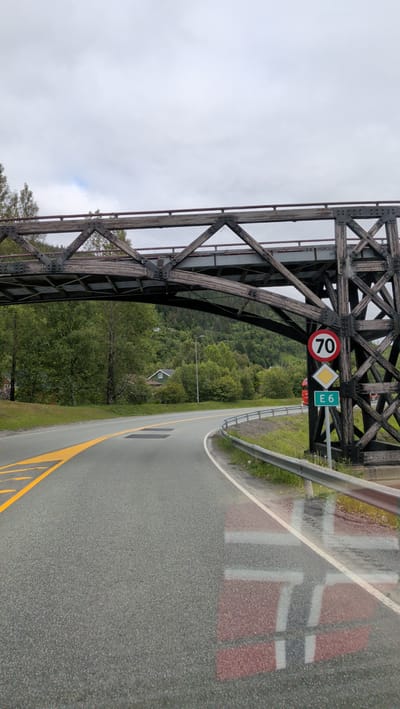The media file captures a view from a vehicle on a wet, grey-skied day, likely in Mosjøen, Norway. The scene is dominated by a sturdy wooden and metal bridge crossing over a paved road. The road curves to the right, with yellow lane markings visible. A metal guardrail runs alongside the road, and patches of grass and vegetation are present.
On the right side of the road, a cluster of traffic signs is mounted on a dark, structural support of the bridge. These signs include a circular speed limit sign indicating "70," a diamond-shaped warning sign, and a rectangular green sign with the text "E6."
In the background, a lush, green, tree-covered hillside rises, with a small house visible nestled among the trees. The sky is overcast and the road surface appears damp, suggesting recent or ongoing rain. The overall mood is somewhat subdued due to the weather. A reflection of what appears to be a large red and white symbol, possibly a Norwegian flag or part of a road marking, is visible on the wet asphalt in the foreground. No people are visible in the image.