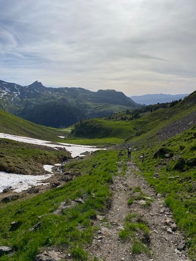 The image shows a mountain landscape in Axalp, Switzerland. A person is walking on a dirt path in the foreground, which is flanked by green grass and rocks. Snow patches are visible on the left side of the path, and a small stream flows nearby. In the background, rolling green hills and distant mountains are visible under a partly cloudy sky. The scene depicts a hiking trail in a natural, mountainous environment.