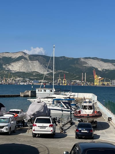 The image captures a sunny day at a marina in Maiorovskii, Russia, with a prominent mountainous landscape in the background. The foreground features a paved area with several vehicles and boats. A white SUV is parked on the right, facing away from the camera, with a boat trailer behind it. To its left, a smaller white boat is loaded onto a trailer, with another white SUV parked beside it. Further left, parts of other boats are visible, some covered with tarpaulins.

In the middle ground, several boats are docked or moored in the blue water. One white sailboat with a tall mast stands out. A small, light-colored building with windows is situated on a concrete structure extending into the water.

The background displays a bustling port area with numerous cranes, industrial structures, and a quarry on the side of a large mountain. Tall chimneys with red and white stripes are visible, along with yellow industrial cranes. The sky is a clear, bright blue with a few scattered white clouds.